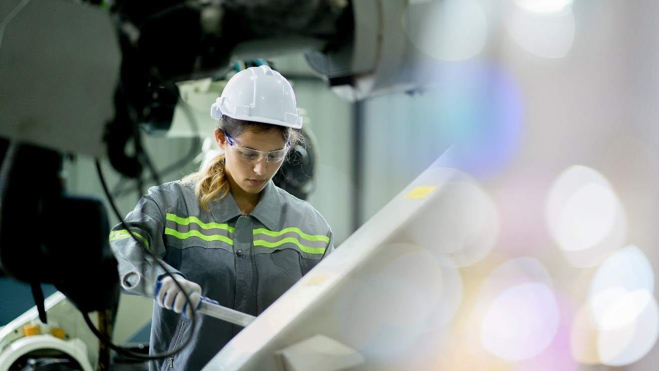 A woman in hard hat, goggles, and work shirt surrounded by machines holds a tool and looks down to work on something with it. 