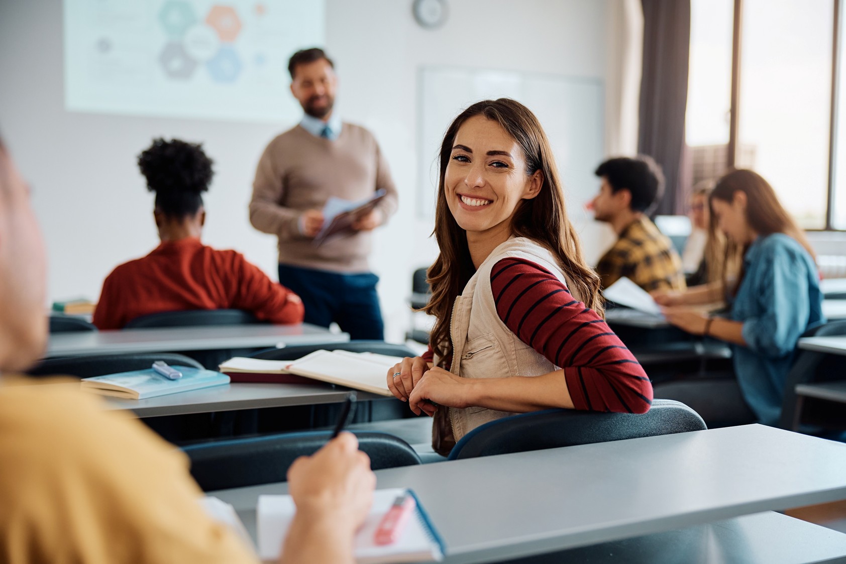 A smiling woman sitting in a classroom turns around while a teacher behind her stands at the front of the room. 