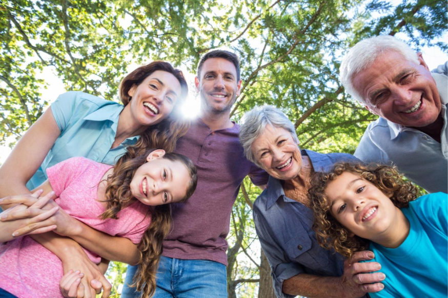 A smiling family of two children, their parents, and their grandparents pictured from below looking down at the ground with trees overhead. 