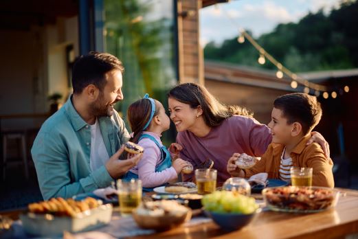A happy dad, daughter, mom, and son sit outside at a table eating, the daughter and mom are touching their noses together. 