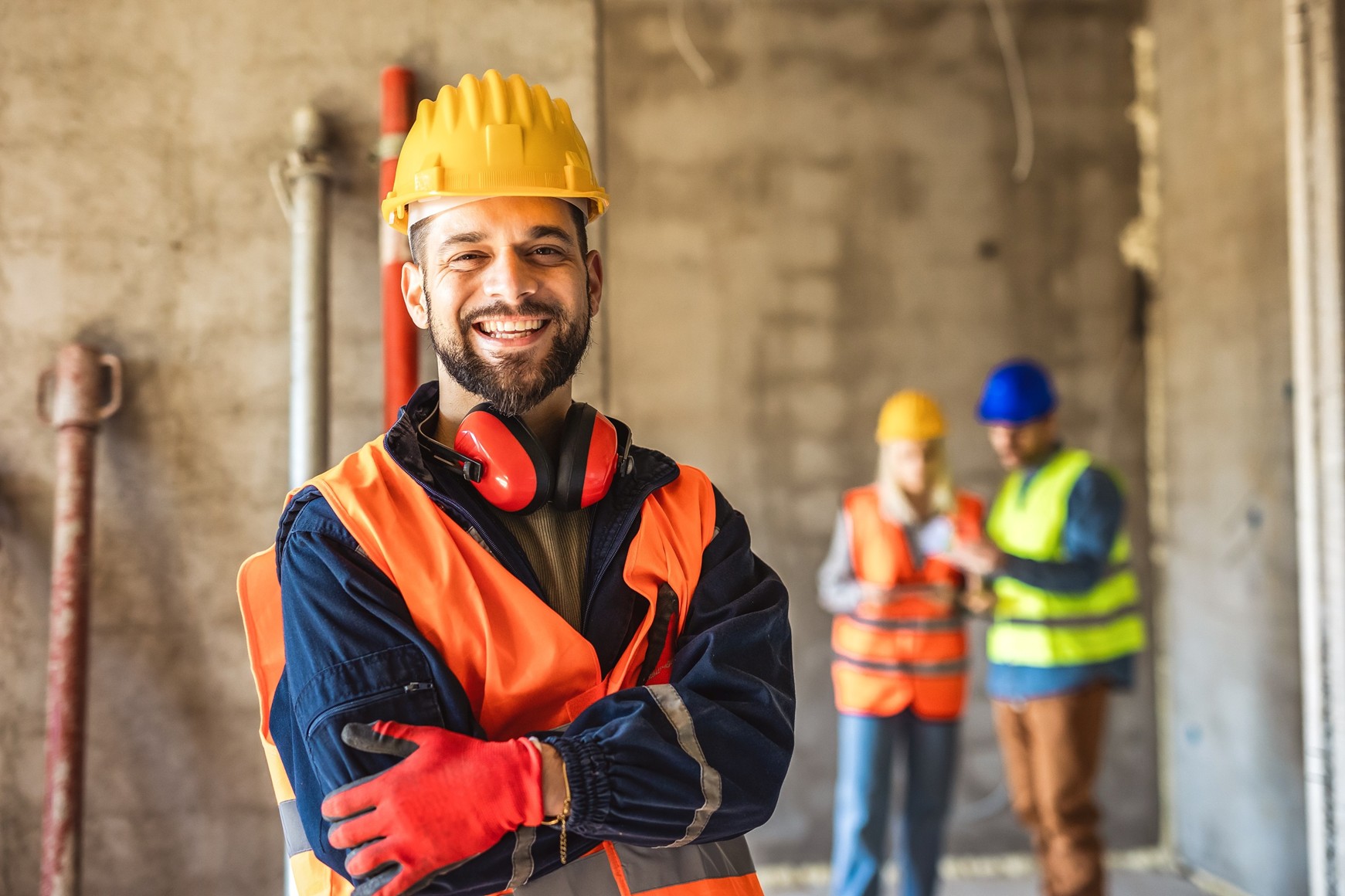A smiling man in a hard hat stands inside an unfinished concrete room with two more construction workers in the background.