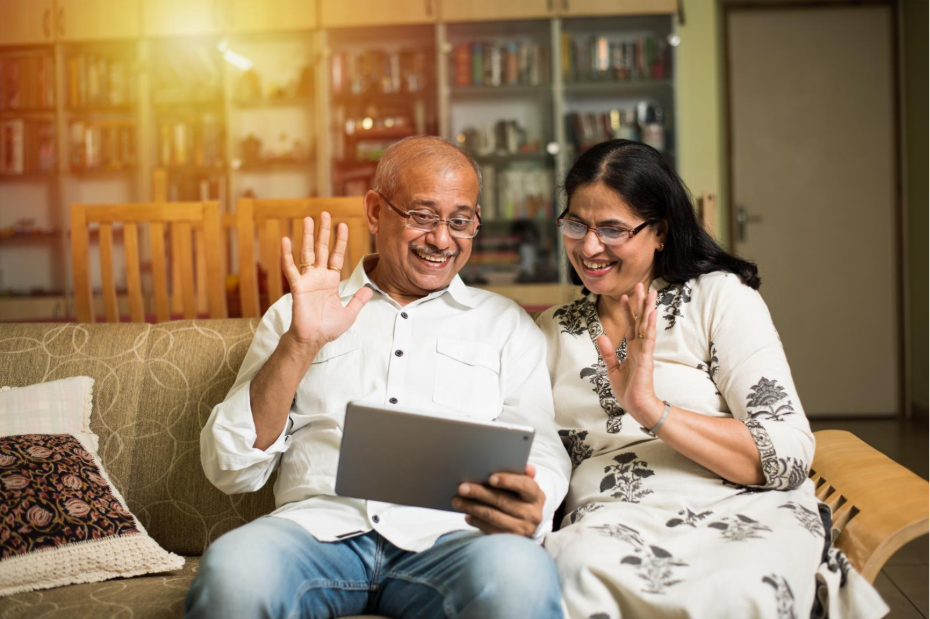 An older Indian couple sits on a couch smiling and waving at a tablet that the man is holding in front of them.