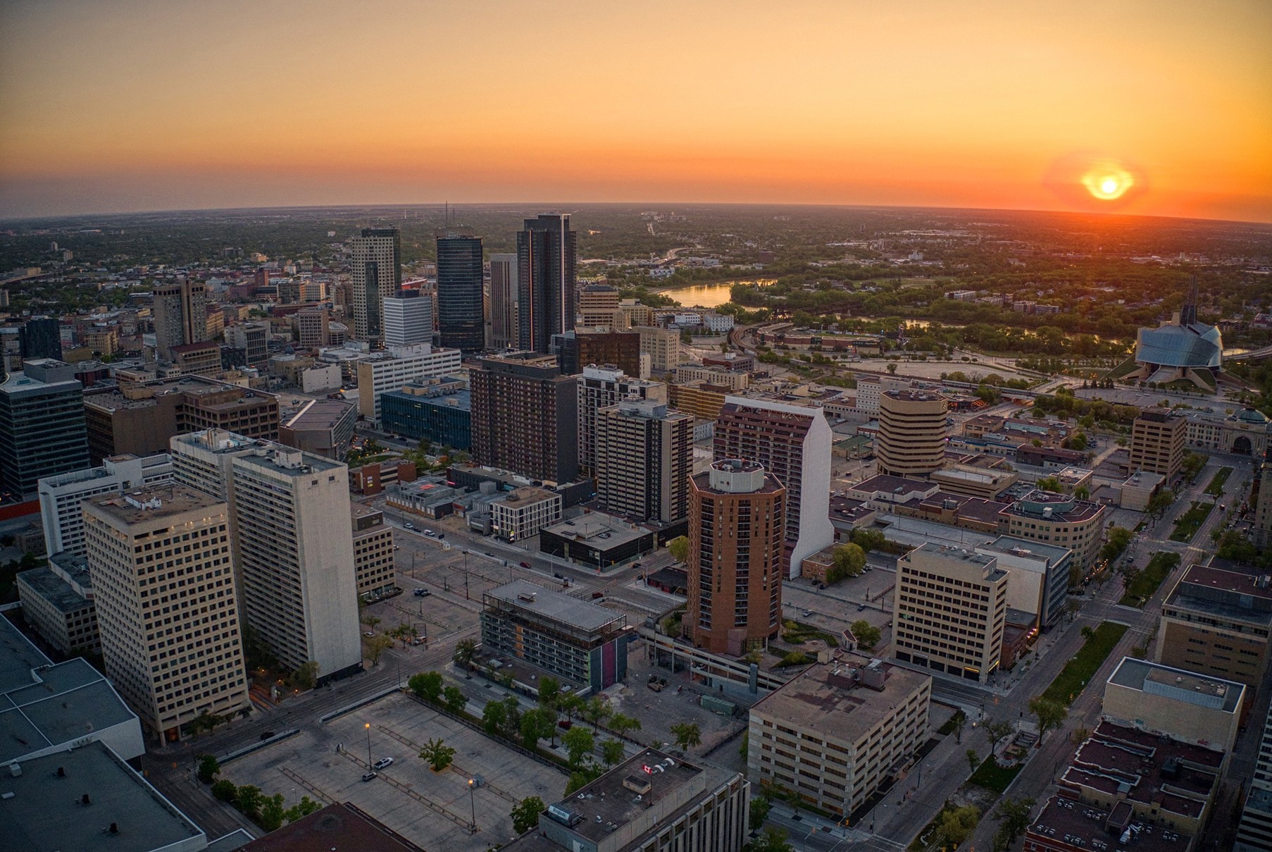 An aerial view of city buildings and streets in Winnipeg, Manitoba, with a setting sun in the background. 