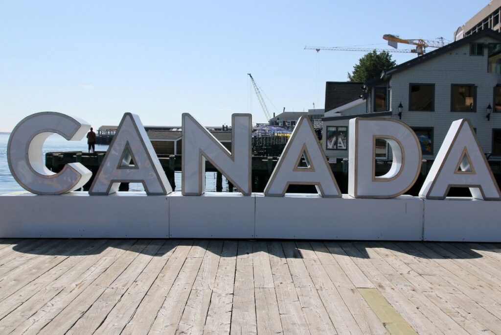 A large sign on a dock with people-sized letters that says “Canada” and a wharf in the background.