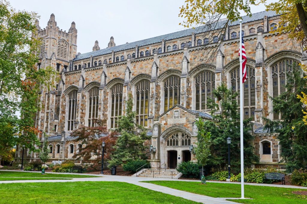 A gothic-style building on the University of Michigan campus with a flagpole with an American flag in front of it. 