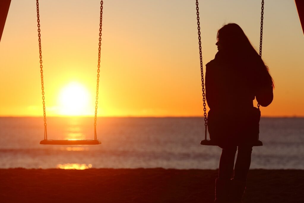 A swing set with two swings, with a woman in silhouette sitting on the right swing in front of the ocean and sunset.