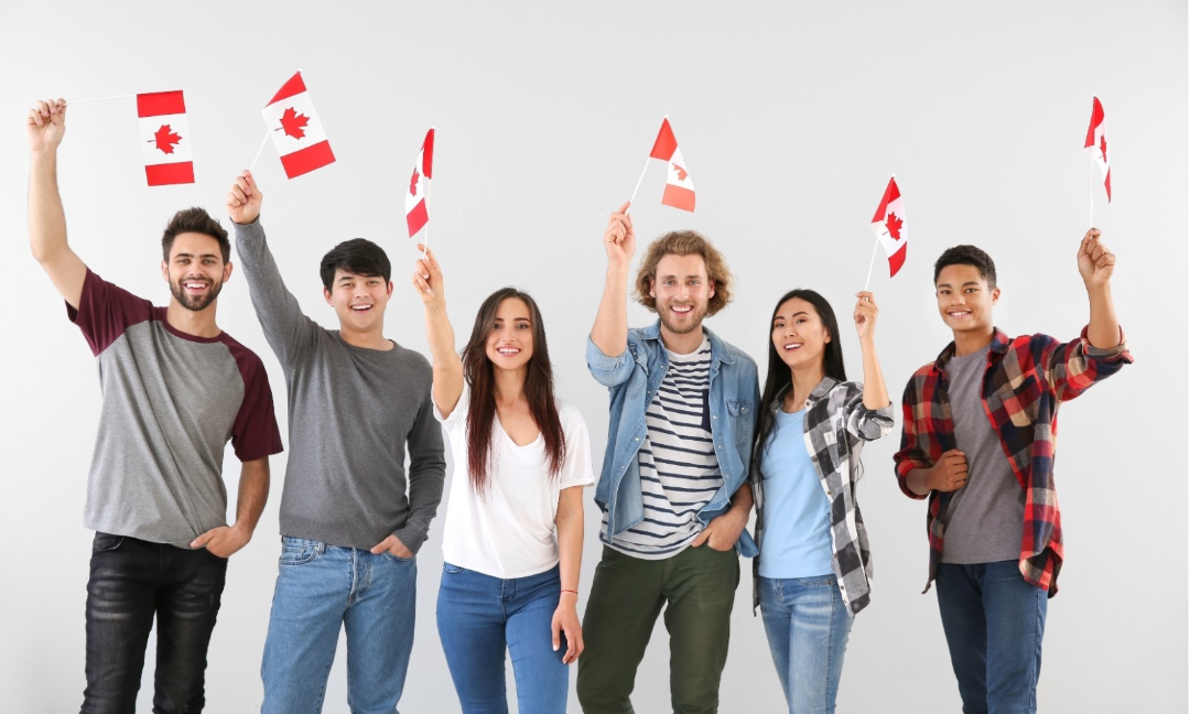 Six young men and women participating in work or study permits, standing together and holding up small Canadian flags. 