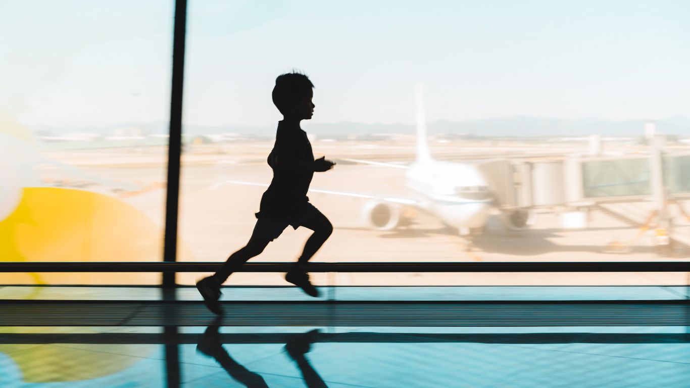  silhouette of a little boy running in an airport next to a window with the plane ready to be boarded in the background.