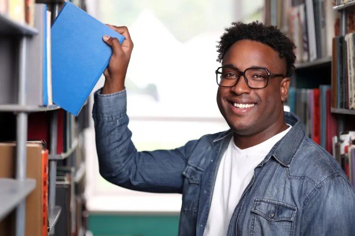 A smiling college-aged man in glasses pulls a blue book from a shelf in a library or bookstore.