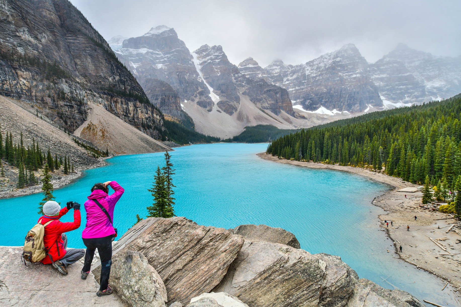 Two people taking a picture of a lake and mountains.