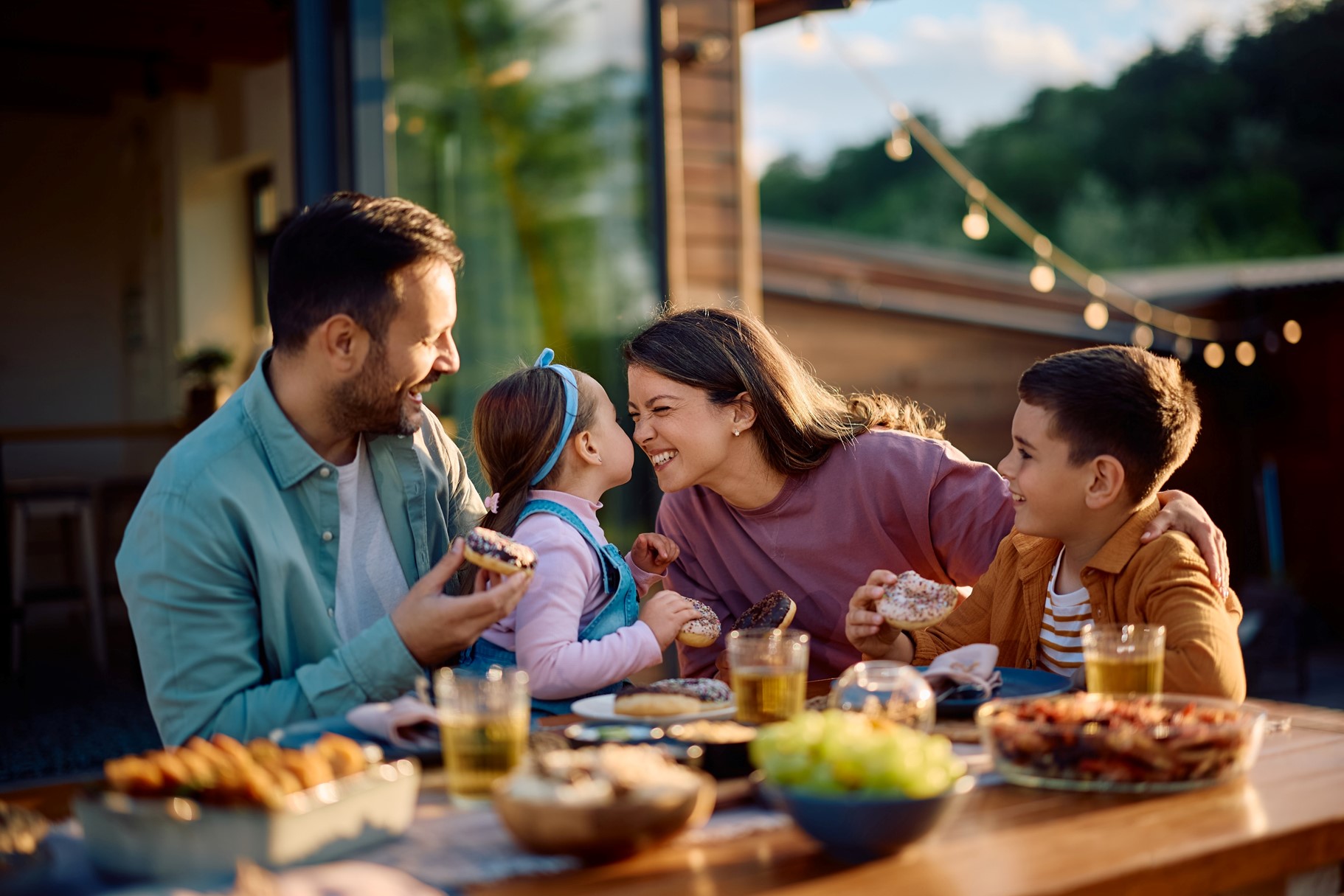 Picture1 A happy dad, daughter, mom, and son sit outside at a table eating, the daughter and mom are touching their noses together.
