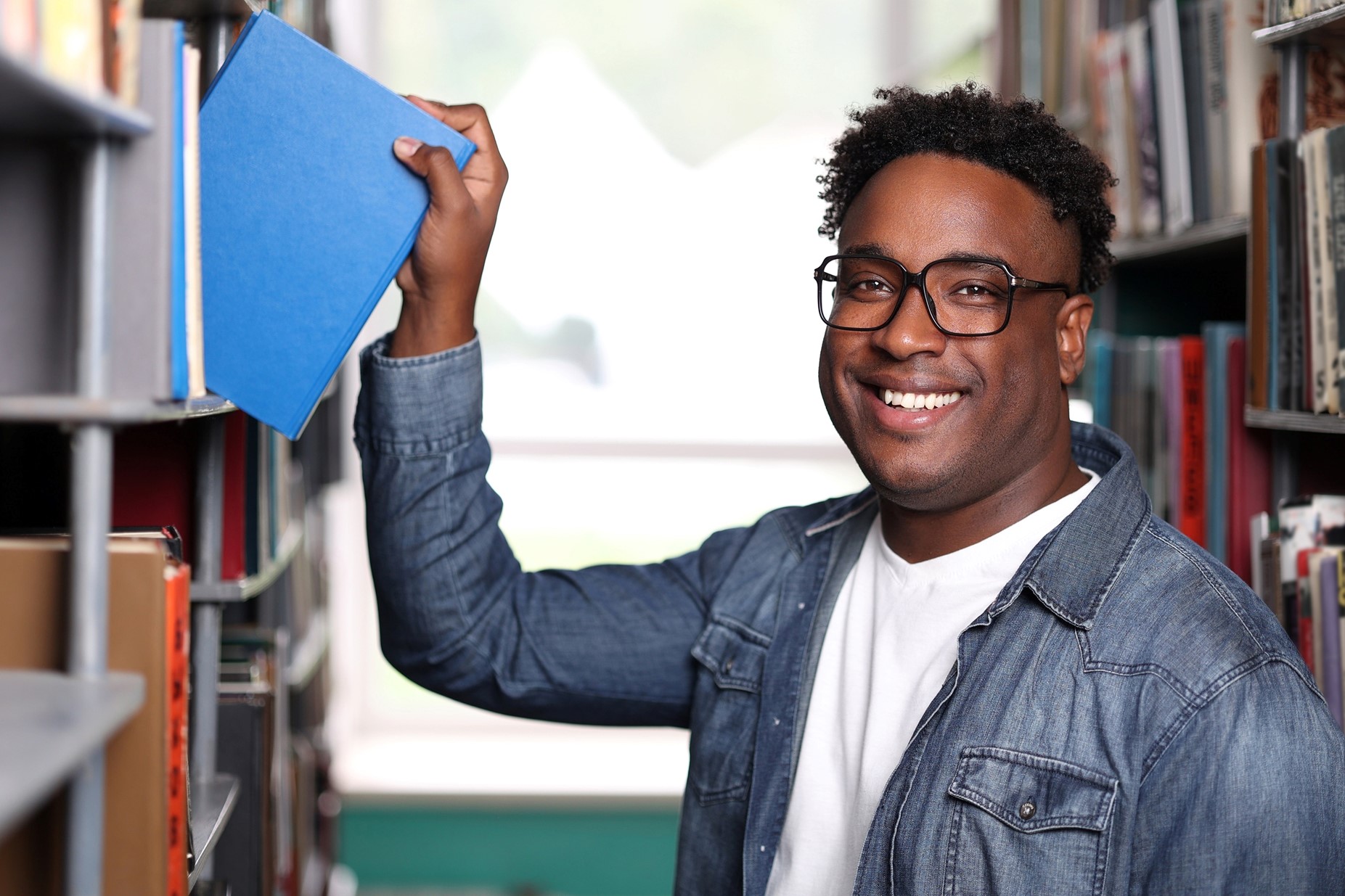 work-and-study-permits A smiling college-aged man in glasses pulls a blue book from a shelf in a library or bookstore.
